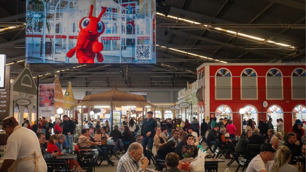 Vista ampla e movimentada da Praça de Alimentação da Fenadoce, em Pelotas. O pavilhão possui arquitetura interna que simula fachadas de casas históricas coloridas. Centenas de pessoas estão sentadas em mesas de madeira, conversando e comendo. No alto, um grande telão exibe o "Formiguinha", mascote oficial da feira, um personagem vermelho sorridente. A atmosfera é vibrante, típica de um grande evento cultural e gastronômico.