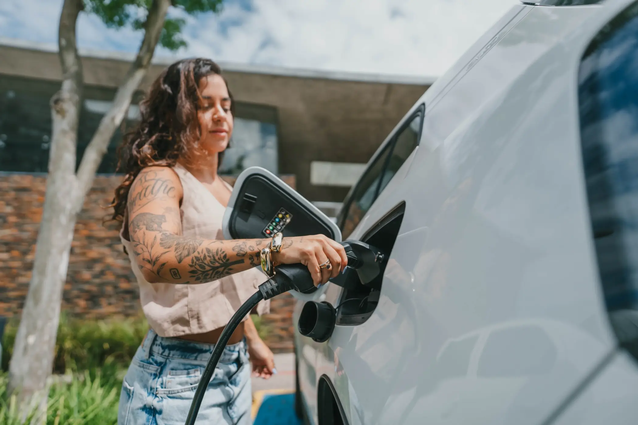pelotas para famílias: Uma mulher com tatuagens de manga inteira no braço direito, vestindo uma blusa bege e calças jeans, está segurando a alça de um cabo de carregamento de carro elétrico (EV) e conectando-o à porta de carregamento aberta de um carro elétrico branco. A porta de carregamento aberta revela luzes de status coloridas. O fundo apresenta uma área externa moderna com uma parede de pedra texturizada, vegetação e um prédio de concreto e vidro.
