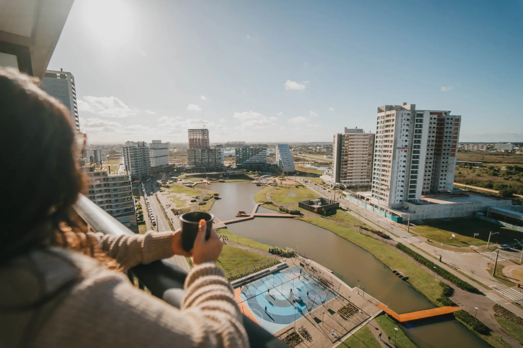 como escolher bairro em pelotas: Vista panorâmica de ângulo alto a partir da varanda de um apartamento, onde aparece em primeiro plano o ombro de uma pessoa segurando uma caneca preta. A visão foca em um bairro planejado moderno com um grande lago artificial sinuoso cortado por passarelas de madeira. Abaixo, destaca-se uma quadra poliesportiva pintada em azul e laranja. Ao redor do lago, erguem-se várias torres de edifícios residenciais contemporâneos, alguns já concluídos e outros em fase de construção estrutural, sob um céu azul ensolarado.