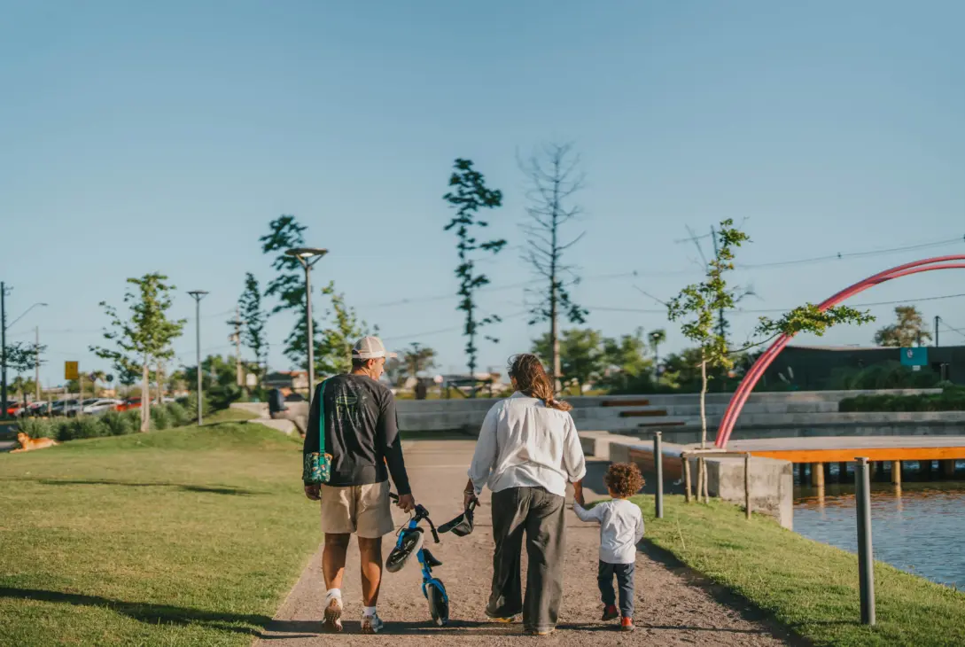 Visão de costas de uma família caminhando por um caminho de cascalho em um parque residencial planejado ao ar livre em um dia ensolarado. O pai, vestindo boné, camiseta preta de manga longa e bermuda clara, caminha carregando uma pequena bicicleta de equilíbrio infantil azul na mão. A mãe, de camisa branca e calça escura, caminha ao lado dele de mãos dadas com um menino pequeno de camiseta branca e calça azul. À direita, a paisagem exibe um lago calmo com um deck de madeira e uma grande estrutura decorativa em formato de arco cor-de-rosa, cercada por gramado e árvores jovens sob um céu azul limpo.