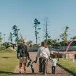 Visão de costas de uma família caminhando por um caminho de cascalho em um parque residencial planejado ao ar livre em um dia ensolarado. O pai, vestindo boné, camiseta preta de manga longa e bermuda clara, caminha carregando uma pequena bicicleta de equilíbrio infantil azul na mão. A mãe, de camisa branca e calça escura, caminha ao lado dele de mãos dadas com um menino pequeno de camiseta branca e calça azul. À direita, a paisagem exibe um lago calmo com um deck de madeira e uma grande estrutura decorativa em formato de arco cor-de-rosa, cercada por gramado e árvores jovens sob um céu azul limpo.