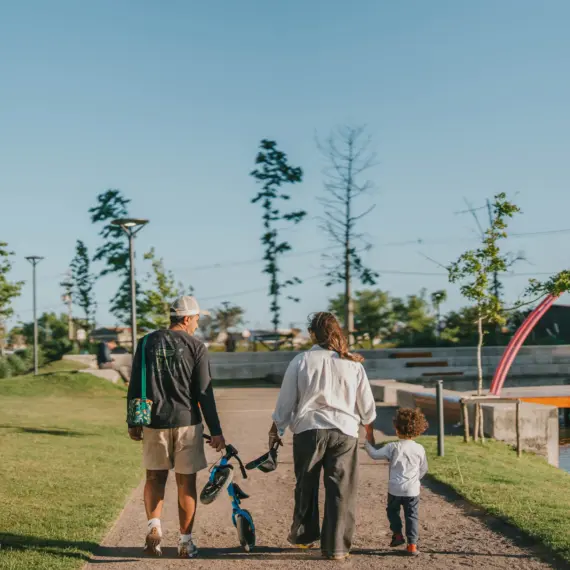 Visão de costas de uma família caminhando por um caminho de cascalho em um parque residencial planejado ao ar livre em um dia ensolarado. O pai, vestindo boné, camiseta preta de manga longa e bermuda clara, caminha carregando uma pequena bicicleta de equilíbrio infantil azul na mão. A mãe, de camisa branca e calça escura, caminha ao lado dele de mãos dadas com um menino pequeno de camiseta branca e calça azul. À direita, a paisagem exibe um lago calmo com um deck de madeira e uma grande estrutura decorativa em formato de arco cor-de-rosa, cercada por gramado e árvores jovens sob um céu azul limpo.