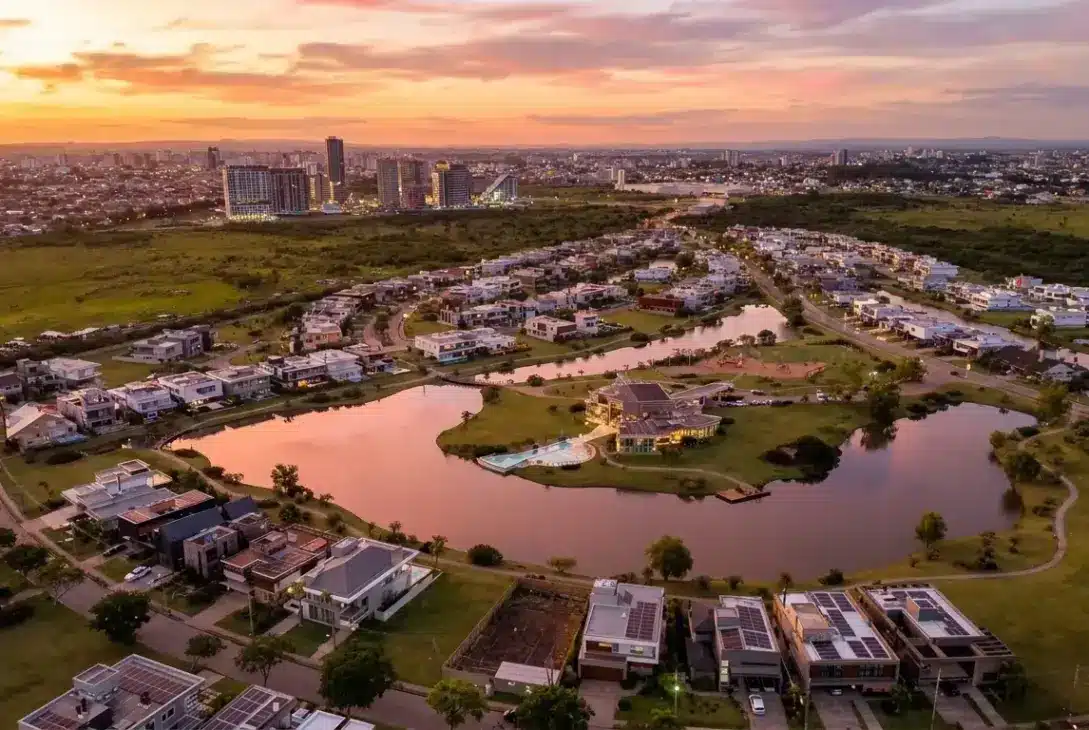 Vista aérea panorâmica de um condomínio residencial de alto padrão planejado em torno de um grande lago artificial com traçado sinuoso. As casas ao redor da água possuem arquitetura contemporânea, telhados planos e muitas contam com placas de energia solar. Áreas gramadas e caminhos contornam toda a extensão do lago. Ao fundo, uma linha de horizonte exibe um denso conjunto de arranha-céus sob um céu com cores vibrantes de laranja, rosa e roxo durante o pôr do sol, contrastando com áreas verdes ainda não urbanizadas nas laterais.
