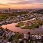 Vista aérea panorâmica de um condomínio residencial de alto padrão planejado em torno de um grande lago artificial com traçado sinuoso. As casas ao redor da água possuem arquitetura contemporânea, telhados planos e muitas contam com placas de energia solar. Áreas gramadas e caminhos contornam toda a extensão do lago. Ao fundo, uma linha de horizonte exibe um denso conjunto de arranha-céus sob um céu com cores vibrantes de laranja, rosa e roxo durante o pôr do sol, contrastando com áreas verdes ainda não urbanizadas nas laterais.