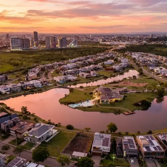 Vista aérea panorâmica de um condomínio residencial de alto padrão planejado em torno de um grande lago artificial com traçado sinuoso. As casas ao redor da água possuem arquitetura contemporânea, telhados planos e muitas contam com placas de energia solar. Áreas gramadas e caminhos contornam toda a extensão do lago. Ao fundo, uma linha de horizonte exibe um denso conjunto de arranha-céus sob um céu com cores vibrantes de laranja, rosa e roxo durante o pôr do sol, contrastando com áreas verdes ainda não urbanizadas nas laterais.
