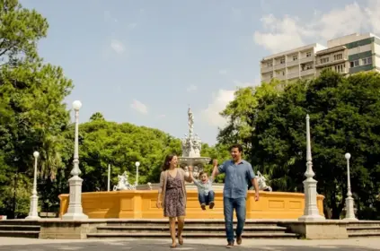 Uma família sorridente passeando em um dia ensolarado pela praça central de Pelotas. Um homem de camisa azul e uma mulher de vestido estampado seguram um menino pequeno pelas mãos, balançando-o no ar enquanto caminham. Ao fundo, ergue-se a icônica Fonte das Nereidas, um grande chafariz ornamental branco cercado por postes de luz clássicos e uma densa vegetação de árvores integradas à praça. Um prédio residencial alto se projeta discretamente acima das árvores no lado direito.