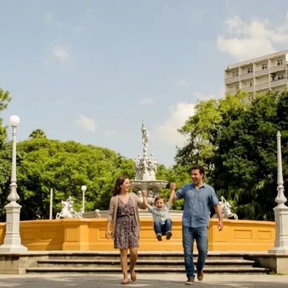Uma família sorridente passeando em um dia ensolarado pela praça central de Pelotas. Um homem de camisa azul e uma mulher de vestido estampado seguram um menino pequeno pelas mãos, balançando-o no ar enquanto caminham. Ao fundo, ergue-se a icônica Fonte das Nereidas, um grande chafariz ornamental branco cercado por postes de luz clássicos e uma densa vegetação de árvores integradas à praça. Um prédio residencial alto se projeta discretamente acima das árvores no lado direito.