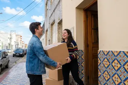 Um jovem de jaqueta jeans e uma jovem de suéter listrado carregam juntos uma grande caixa de papelão na calçada, logo em frente à entrada de um prédio residencial. Ambos estão rindo e se olhando com cumplicidade. A fachada inferior do edifício é decorada com azulejos antigos em tons de azul, laranja e amarelo. No chão, há mais caixas de papelão empilhadas perto da porta de madeira. Ao fundo, vê-se uma rua residencial com carros estacionados sob um céu azul claro. (aluguel para estudantes pelotas)