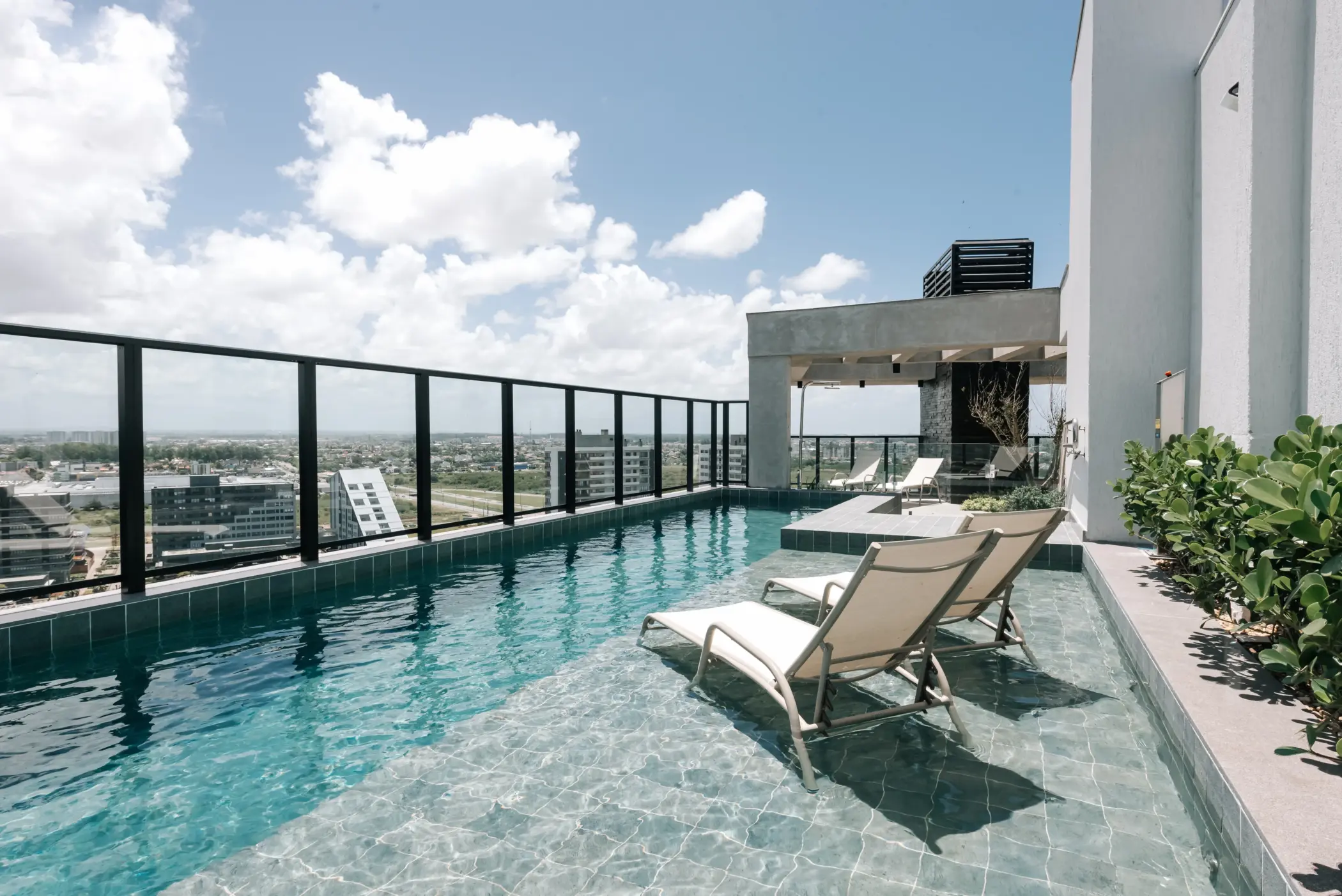 Piscina de borda infinita localizada na cobertura de um edifício residencial moderno. Duas espreguiçadeiras brancas estão posicionadas sobre a prainha rasa da água. O espaço conta com guarda-corpo de vidro voltado para a vista panorâmica da cidade sob céu ensolarado com nuvens.