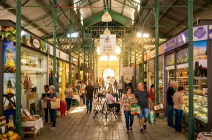 Corredor central de um mercado público histórico caracterizado por colunas e estrutura metálica verde sobre paredes de tijolo à vista. Vários visitantes caminham entre lojas envidraçadas de doces e produtos locais, com um casal em primeiro plano carregando sacolas de compras.