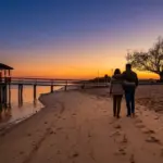 Fotografia vista de costas de um casal caminhando abraçado pela faixa de areia ao entardecer. O sol poente reflete tons quentes de laranja e dourado sobre a água calma, destacando um trapiche de madeira com quiosque e a silhueta de árvores à beira da praia.