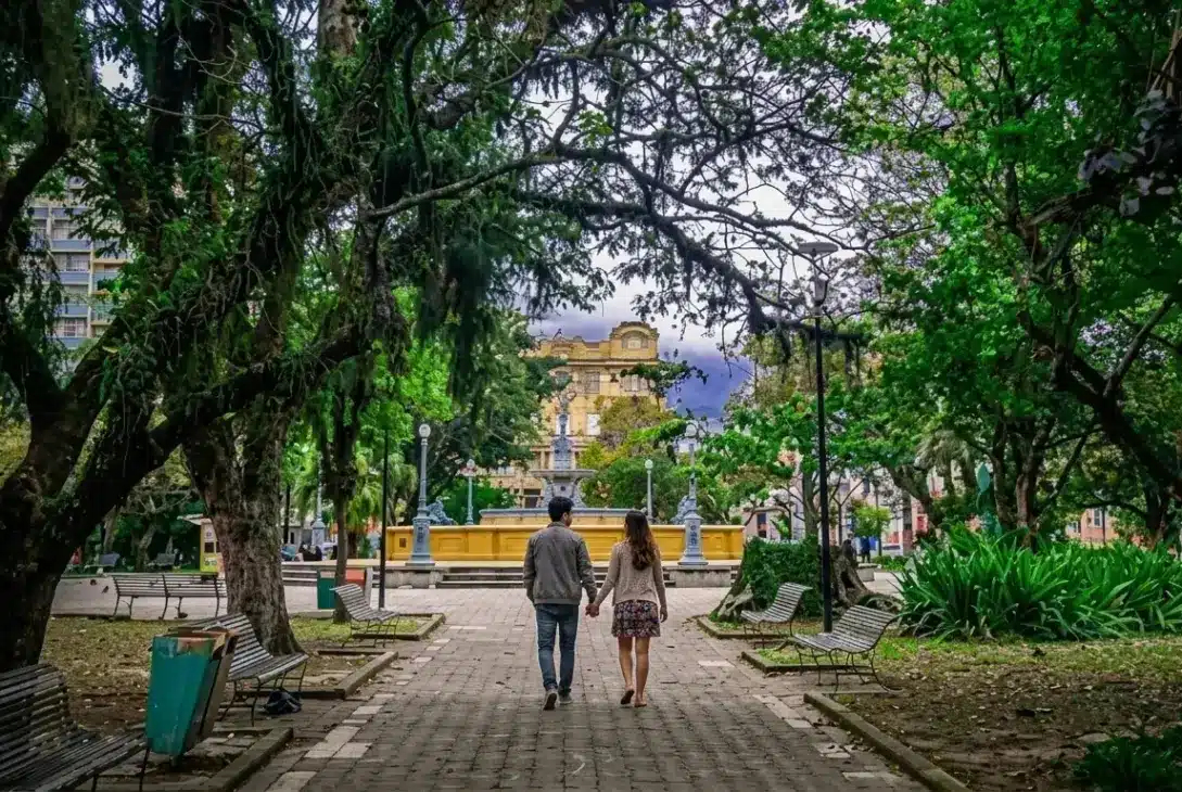 Fotografia vista de costas de um casal caminhando de mãos dadas por uma aladeira pavimentada no centro de uma praça pública bastante arborizada. O caminho ladeado por bancos de madeira e copa de grandes árvores conduz a um chafariz histórico amarelado, com edifícios clássicos ao fundo.