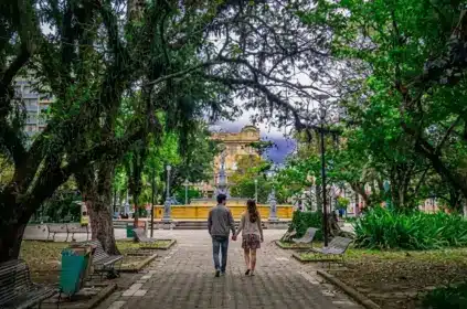 Fotografia vista de costas de um casal caminhando de mãos dadas por uma aladeira pavimentada no centro de uma praça pública bastante arborizada. O caminho ladeado por bancos de madeira e copa de grandes árvores conduz a um chafariz histórico amarelado, com edifícios clássicos ao fundo.