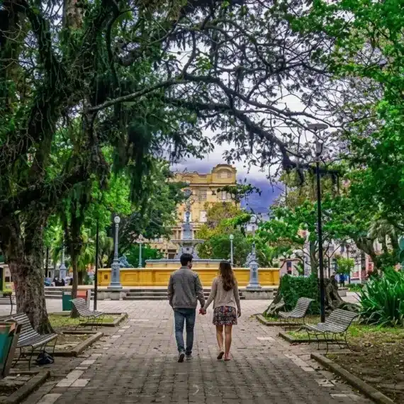 Fotografia vista de costas de um casal caminhando de mãos dadas por uma aladeira pavimentada no centro de uma praça pública bastante arborizada. O caminho ladeado por bancos de madeira e copa de grandes árvores conduz a um chafariz histórico amarelado, com edifícios clássicos ao fundo.