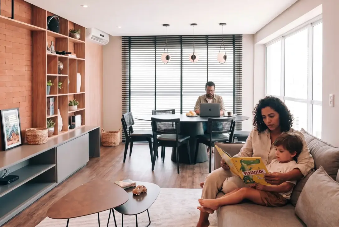 Uma família em uma sala de estar moderna e integrada. Em primeiro plano, uma mulher de cabelo cacheado lê um livro infantil ilustrado para uma criança pequena em seu colo no sofá. Ao fundo, um homem de óculos trabalha em um notebook em uma mesa de jantar redonda, em um ambiente decorado com parede de tijolos aparentes, estantes de madeira e amplas janelas.