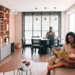 Uma família em uma sala de estar moderna e integrada. Em primeiro plano, uma mulher de cabelo cacheado lê um livro infantil ilustrado para uma criança pequena em seu colo no sofá. Ao fundo, um homem de óculos trabalha em um notebook em uma mesa de jantar redonda, em um ambiente decorado com parede de tijolos aparentes, estantes de madeira e amplas janelas.