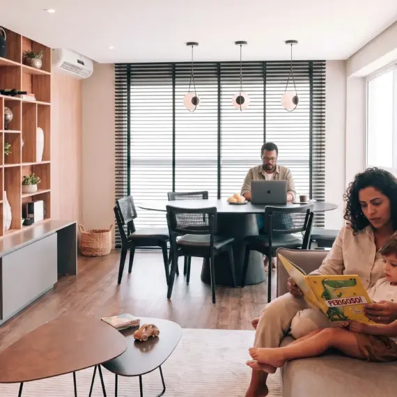 Uma família em uma sala de estar moderna e integrada. Em primeiro plano, uma mulher de cabelo cacheado lê um livro infantil ilustrado para uma criança pequena em seu colo no sofá. Ao fundo, um homem de óculos trabalha em um notebook em uma mesa de jantar redonda, em um ambiente decorado com parede de tijolos aparentes, estantes de madeira e amplas janelas.