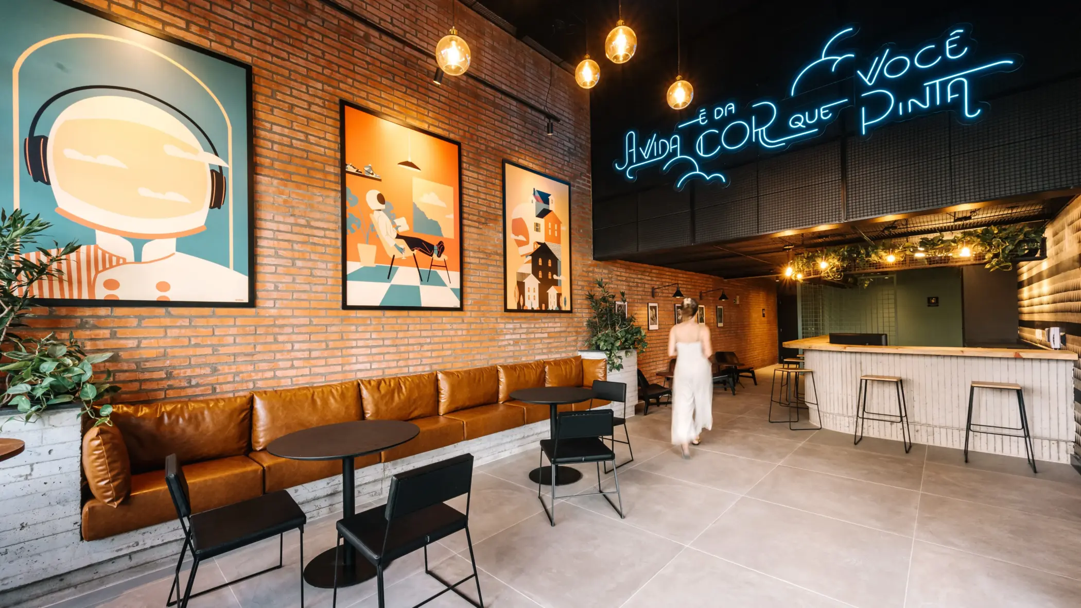 Um espaço de convivência em estilo industrial com parede de tijolos à vista e quadros artísticos emoldurados. O ambiente conta com sofá extenso de couro caramelo, mesas de apoio pretas, lâmpadas pendentes e um letreiro em néon azul com a frase "A VIDA É DA COR QUE VOCÊ PINTA" fixado acima do balcão.