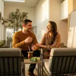 Um casal sorridente e bem-vestido conversando em poltronas sob a iluminação dourada do sol da tarde. Em uma pequena mesa de apoio entre eles, observam-se uma cuia de chimarrão e um pacote de erva-mate, em um espaço aconchegante com painéis de madeira.