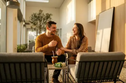 Um casal sorridente e bem-vestido conversando em poltronas sob a iluminação dourada do sol da tarde. Em uma pequena mesa de apoio entre eles, observam-se uma cuia de chimarrão e um pacote de erva-mate, em um espaço aconchegante com painéis de madeira.