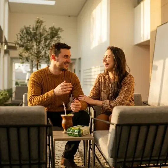Um casal sorridente e bem-vestido conversando em poltronas sob a iluminação dourada do sol da tarde. Em uma pequena mesa de apoio entre eles, observam-se uma cuia de chimarrão e um pacote de erva-mate, em um espaço aconchegante com painéis de madeira.