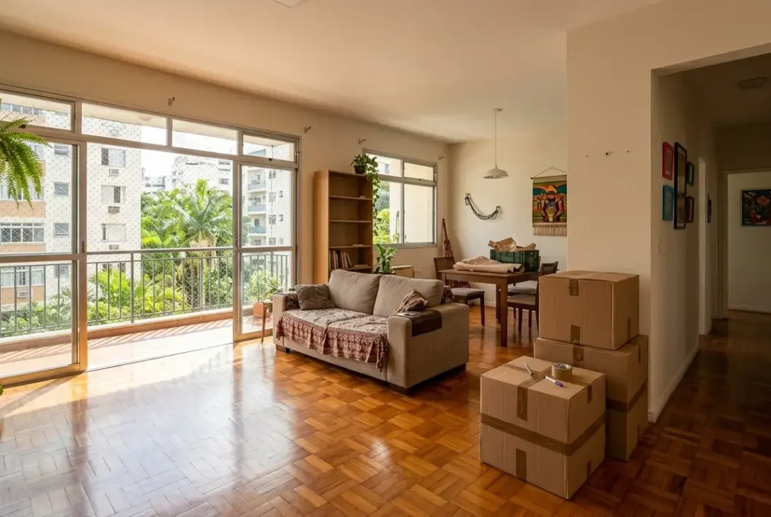 Sala de estar iluminada de um apartamento em processo de mudança, com piso de taco de madeira, sofá e portas de vidro com vista para a varanda, destacando caixas de papelão empilhadas e lacradas com fita em primeiro plano.Sala de estar iluminada de um apartamento em processo de mudança, com piso de taco de madeira, sofá e portas de vidro com vista para a varanda, destacando caixas de papelão empilhadas e lacradas com fita em primeiro plano.