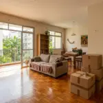 Sala de estar iluminada de um apartamento em processo de mudança, com piso de taco de madeira, sofá e portas de vidro com vista para a varanda, destacando caixas de papelão empilhadas e lacradas com fita em primeiro plano.Sala de estar iluminada de um apartamento em processo de mudança, com piso de taco de madeira, sofá e portas de vidro com vista para a varanda, destacando caixas de papelão empilhadas e lacradas com fita em primeiro plano.
