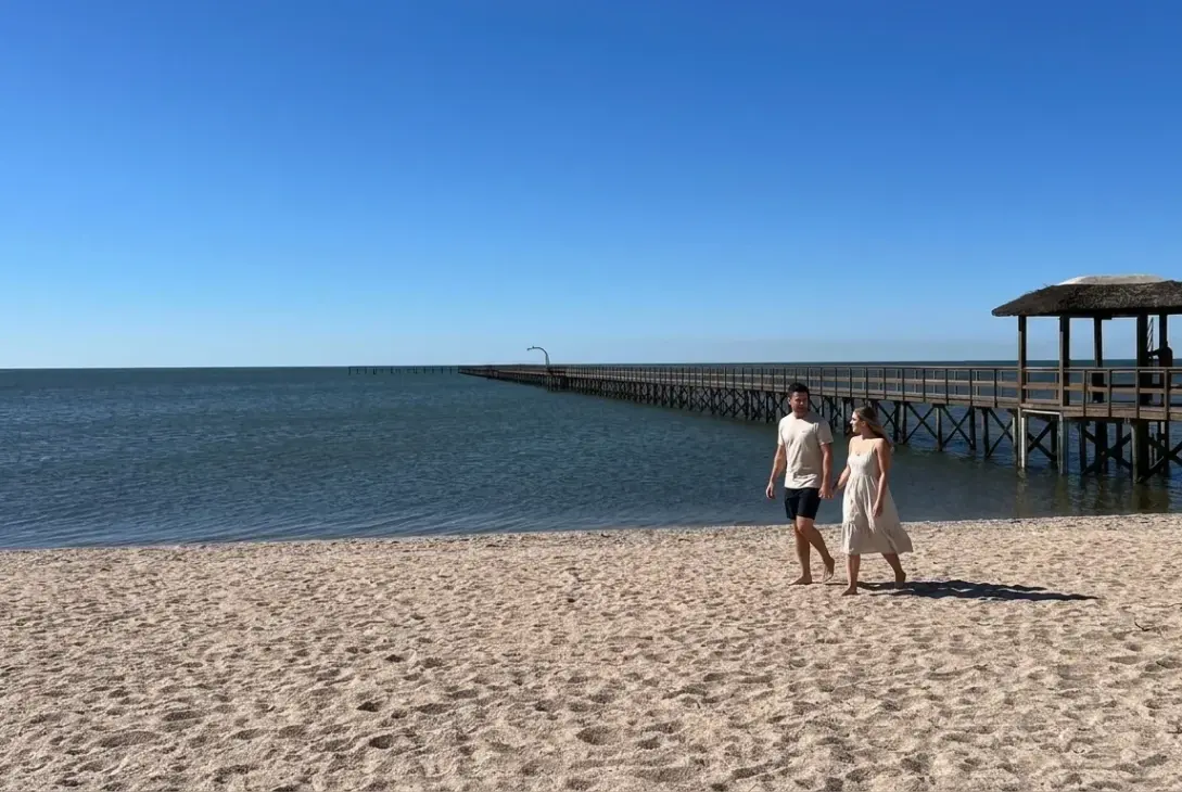 Um casal caminha descalço e de mãos dadas pela areia de uma praia ensolarada, com um longo trapiche de madeira e quiosque de palha avançando sobre a água calma ao fundo sob céu azul.
