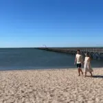 Um casal caminha descalço e de mãos dadas pela areia de uma praia ensolarada, com um longo trapiche de madeira e quiosque de palha avançando sobre a água calma ao fundo sob céu azul.