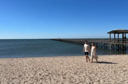 Um casal caminha descalço e de mãos dadas pela areia de uma praia ensolarada, com um longo trapiche de madeira e quiosque de palha avançando sobre a água calma ao fundo sob céu azul.