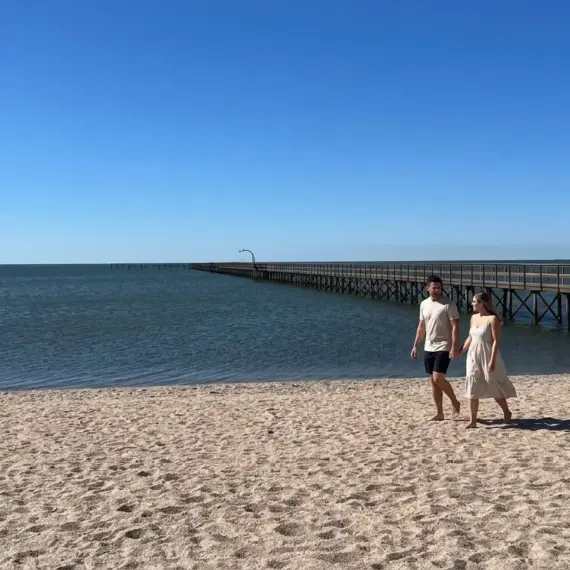 Um casal caminha descalço e de mãos dadas pela areia de uma praia ensolarada, com um longo trapiche de madeira e quiosque de palha avançando sobre a água calma ao fundo sob céu azul.