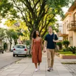 Um casal caminhando de mãos dadas pela calçada de uma rua residencial tranquila e arborizada. A mulher veste um vestido terracota e o homem usa camisa polo marinho com bolsa transversal, observando os prédios ao redor, com destaque para a fachada do "EDIFÍCIO JARDIM DAS FLORES".