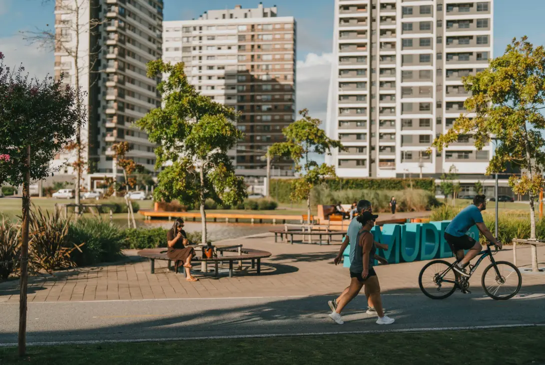 Pessoas aproveitam um parque urbano ensolarado com pista pavimentada, onde um homem anda de bicicleta e um casal caminha em primeiro plano, enquanto uma mulher toma chimarrão sentada em um banco circular ao lado de um lago, com grandes edifícios modernos ao fundo.