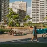 Pessoas aproveitam um parque urbano ensolarado com pista pavimentada, onde um homem anda de bicicleta e um casal caminha em primeiro plano, enquanto uma mulher toma chimarrão sentada em um banco circular ao lado de um lago, com grandes edifícios modernos ao fundo.