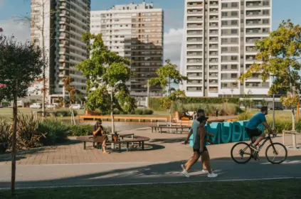 Pessoas aproveitam um parque urbano ensolarado com pista pavimentada, onde um homem anda de bicicleta e um casal caminha em primeiro plano, enquanto uma mulher toma chimarrão sentada em um banco circular ao lado de um lago, com grandes edifícios modernos ao fundo.